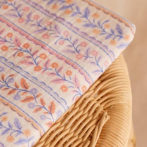 May include: A folded fabric with a floral pattern in shades of pink, orange, and blue on a cream background. The fabric is resting on a woven wicker surface, creating a contrast in textures. The floral design features delicate vines and small flowers.