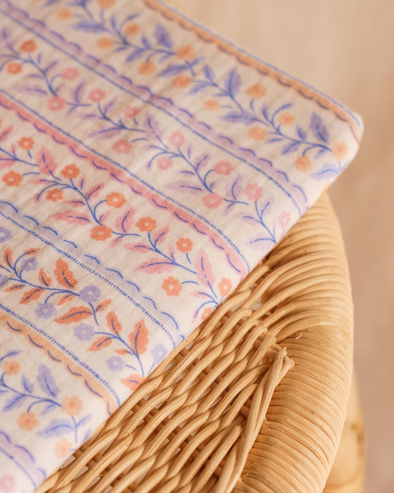 May include: A folded fabric with a floral pattern in shades of pink, orange, and blue on a cream background. The fabric is resting on a woven wicker surface, creating a contrast in textures. The floral design features delicate vines and small flowers.