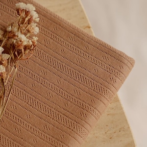 May include: A folded, light brown fabric with a detailed, openwork pattern. The fabric is draped over a light-colored, textured surface. A sprig of dried flowers with white and brown blossoms rests on the fabric.