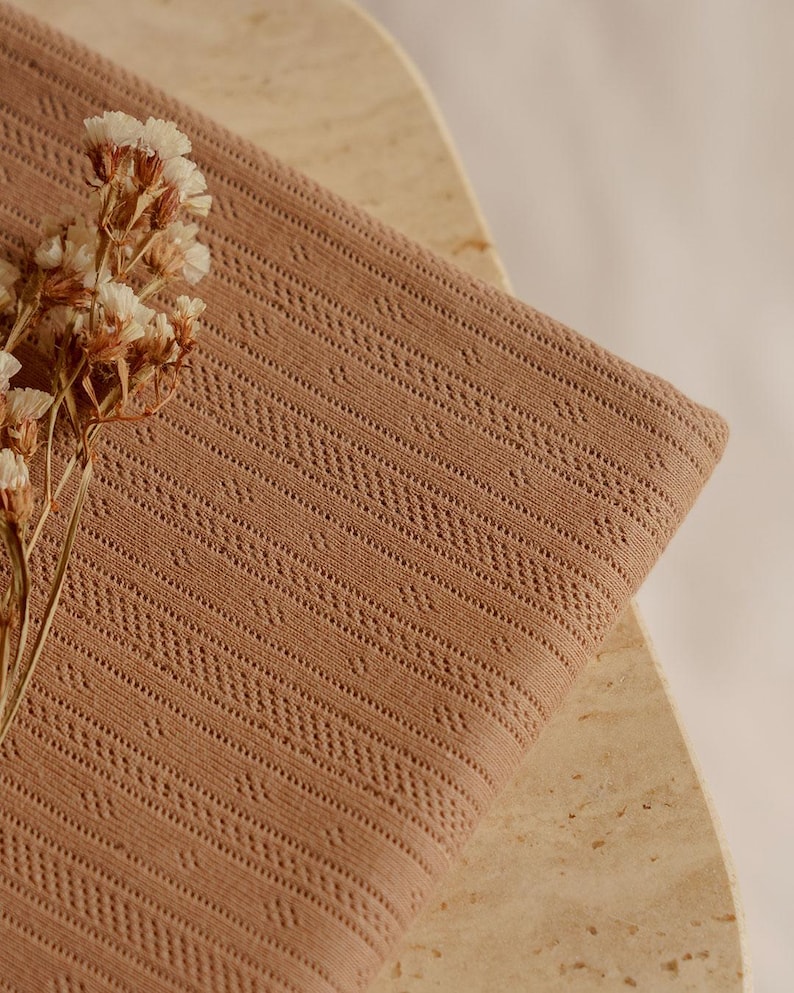May include: A folded, light brown fabric with a detailed, openwork pattern. The fabric is draped over a light-colored, textured surface. A sprig of dried flowers with white and brown blossoms rests on the fabric.