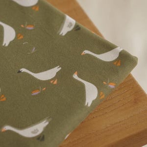 Pu&ograve; includere: Primo piano di un tessuto piegato con un motivo ripetuto di oche bianche su uno sfondo verde tenue. Le oche hanno becchi e zampe arancioni. Piccoli accenti floreali sono sparsi nel design. Il tessuto &egrave; drappeggiato su una superficie di legno.