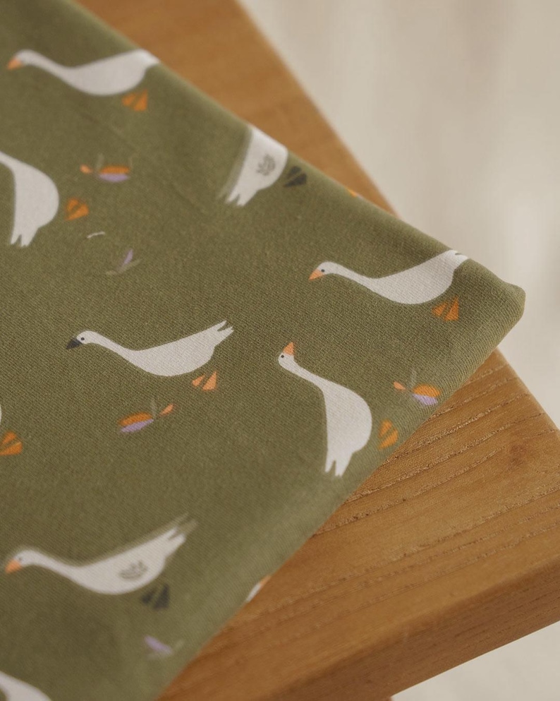 Pu&ograve; includere: Primo piano di un tessuto piegato con un motivo ripetuto di oche bianche su uno sfondo verde tenue. Le oche hanno becchi e zampe arancioni. Piccoli accenti floreali sono sparsi nel design. Il tessuto &egrave; drappeggiato su una superficie di legno.