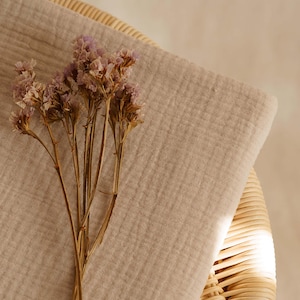 Op de afbeelding: Een beige mousseline stof is over een rieten mand gedrapeerd, met een bosje gedroogde paarse bloemen op de voorgrond. De stof heeft een getextureerde uitstraling, wat een zachte, natuurlijke esthetiek cre&euml;ert.