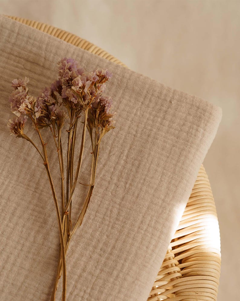 Op de afbeelding: Een beige mousseline stof is over een rieten mand gedrapeerd, met een bosje gedroogde paarse bloemen op de voorgrond. De stof heeft een getextureerde uitstraling, wat een zachte, natuurlijke esthetiek cre&euml;ert.