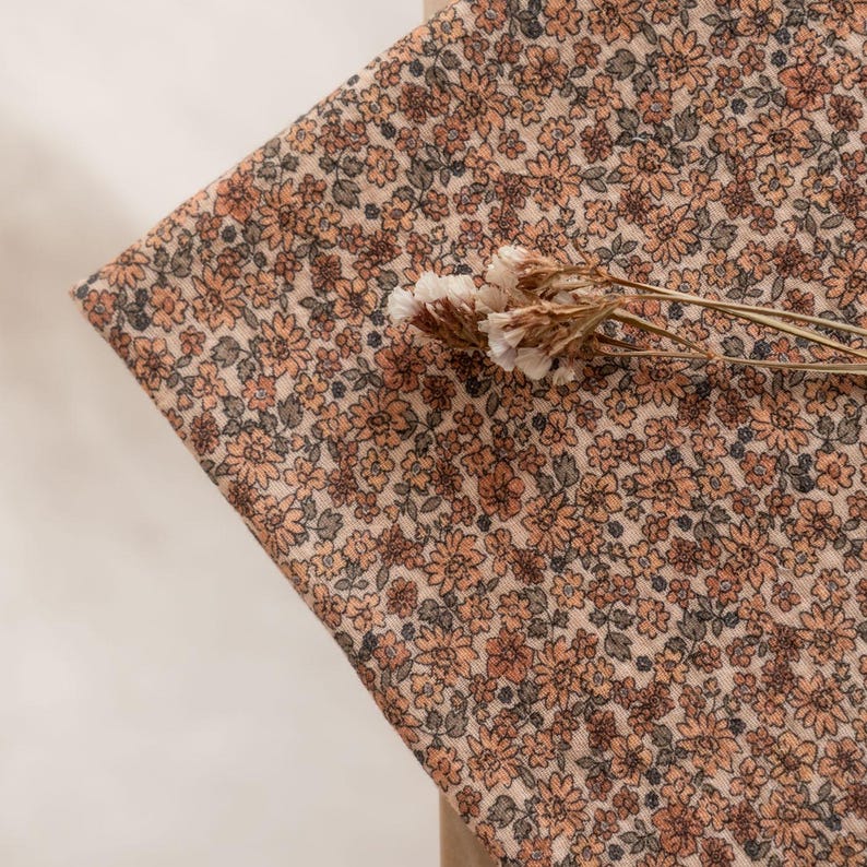 May include: A close-up of a beige fabric with a small-scale floral print in shades of brown and peach. The fabric is textured and has a subtle sheen.