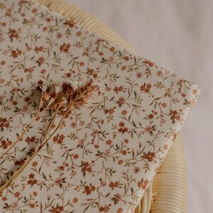 May include: A folded piece of fabric with a floral pattern in shades of brown and pink on an off-white background. The fabric is resting on a woven chair. Dried flowers are placed on top of the fabric.