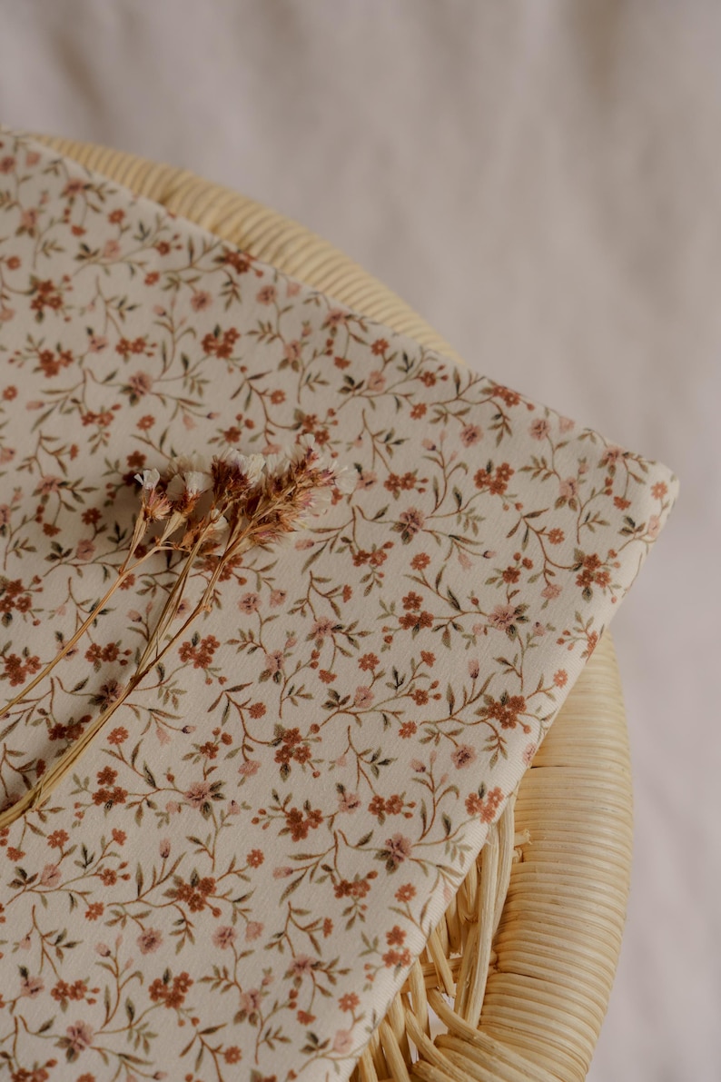 May include: A folded piece of fabric with a floral pattern in shades of brown and pink on an off-white background. The fabric is resting on a woven chair. Dried flowers are placed on top of the fabric.