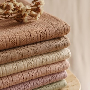 May include: A stack of six folded textiles in varying shades of brown, beige, pink, and green. Each textile has a ribbed texture with a delicate, openwork pattern. A sprig of dried flowers rests on top of the stack, adding a natural element.
