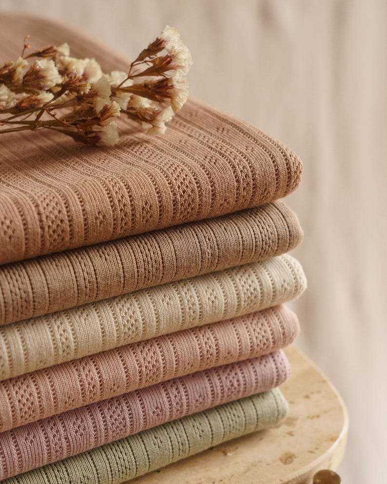 May include: A stack of six folded textiles in varying shades of brown, beige, pink, and green. Each textile has a ribbed texture with a delicate, openwork pattern. A sprig of dried flowers rests on top of the stack, adding a natural element.
