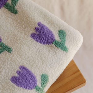 May include: A close-up of a white, textured fabric featuring a repeating pattern of purple tulip flowers with green stems and leaves. The fabric is resting on a small, light-colored wooden stool.