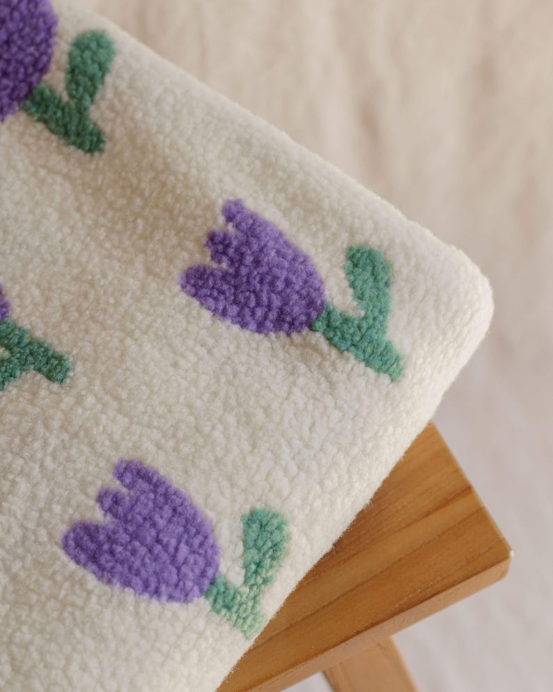 May include: A close-up of a white, textured fabric featuring a repeating pattern of purple tulip flowers with green stems and leaves. The fabric is resting on a small, light-colored wooden stool.