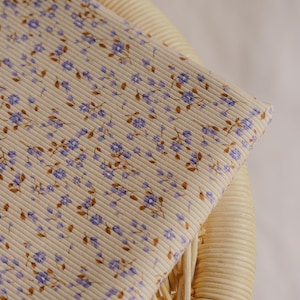 May include: A folded piece of beige fabric with a ribbed texture and a floral pattern. The pattern features small purple flowers and brown stems. The fabric is resting on a woven wicker chair.