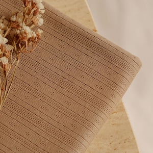 May include: A folded piece of tan fabric with a textured, dotted pattern. The fabric is draped over a light-colored, textured surface. Dried flowers with small, white blossoms are placed on the fabric, adding a natural element to the composition.