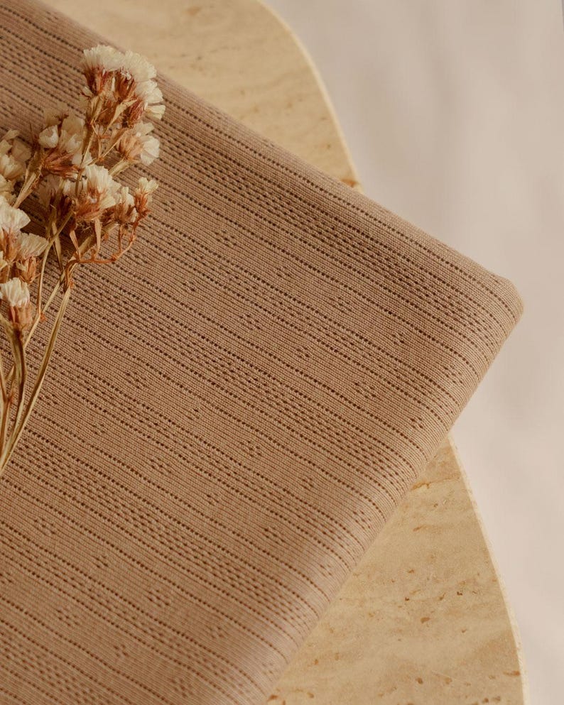 May include: A folded piece of tan fabric with a textured, dotted pattern. The fabric is draped over a light-colored, textured surface. Dried flowers with small, white blossoms are placed on the fabric, adding a natural element to the composition.