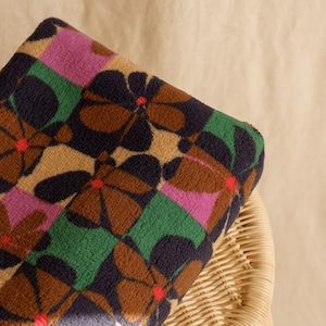 May include: A folded blanket with a repeating floral pattern. The flowers are brown with navy blue petals and red centers, set against a tan background. The pattern includes green and pink squares. The blanket is resting on a wicker chair.