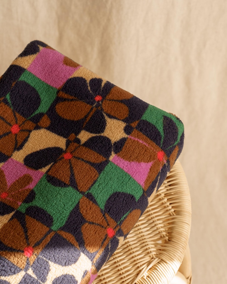 May include: A folded blanket with a repeating floral pattern. The flowers are brown with navy blue petals and red centers, set against a tan background. The pattern includes green and pink squares. The blanket is resting on a wicker chair.
