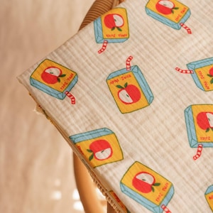 May include: A cream-coloured baby blanket featuring a repeating pattern of apple juice cartons. Each carton is yellow with a red apple graphic, blue trim, and a red and white striped straw. The blanket is draped over a wicker chair.