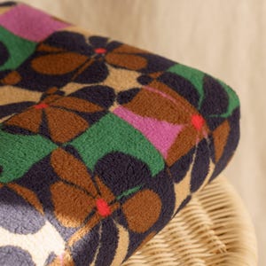 Op de afbeelding: Een opgerolde deken met een herhalend bloemenpatroon. Het ontwerp bevat grote bloemen in bruin, marine, groen, roze en beige. De deken ligt op een gevlochten rieten stoel.