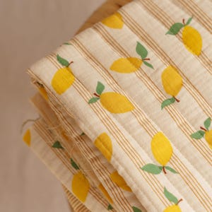 Peut inclure: Tissu plié avec un motif répété de citrons jaunes et de feuilles vertes sur un fond crème à rayures marron verticales. Le tissu semble être matelassé.