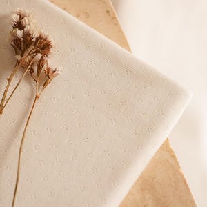 Op de afbeelding: Een close-up van een witte stof met een subtiel textuurpatroon. De stof is gevouwen en heeft een delicate, bijna doorschijnende kwaliteit. Een takje gedroogde bloemen ligt op de stof.
