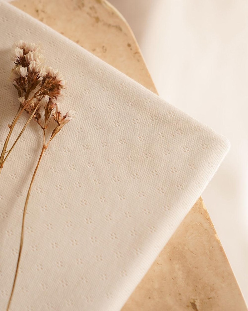 Op de afbeelding: Een close-up van een witte stof met een subtiel textuurpatroon. De stof is gevouwen en heeft een delicate, bijna doorschijnende kwaliteit. Een takje gedroogde bloemen ligt op de stof.