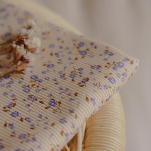 May include: Close-up of a beige fabric with a ribbed texture, adorned with small purple floral patterns and brown leaves. The fabric is folded over a woven wicker surface, with dried flowers in the corner. The overall theme is floral fabric.