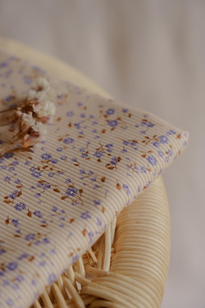 May include: Close-up of a beige fabric with a ribbed texture, adorned with small purple floral patterns and brown leaves. The fabric is folded over a woven wicker surface, with dried flowers in the corner. The overall theme is floral fabric.
