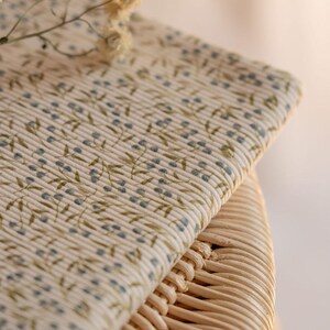 May include: Close-up of folded cream fabric with a floral design. The pattern shows small blue berries and green leaves on stems. The fabric rests on a woven wicker surface. The overall aesthetic is soft and natural.