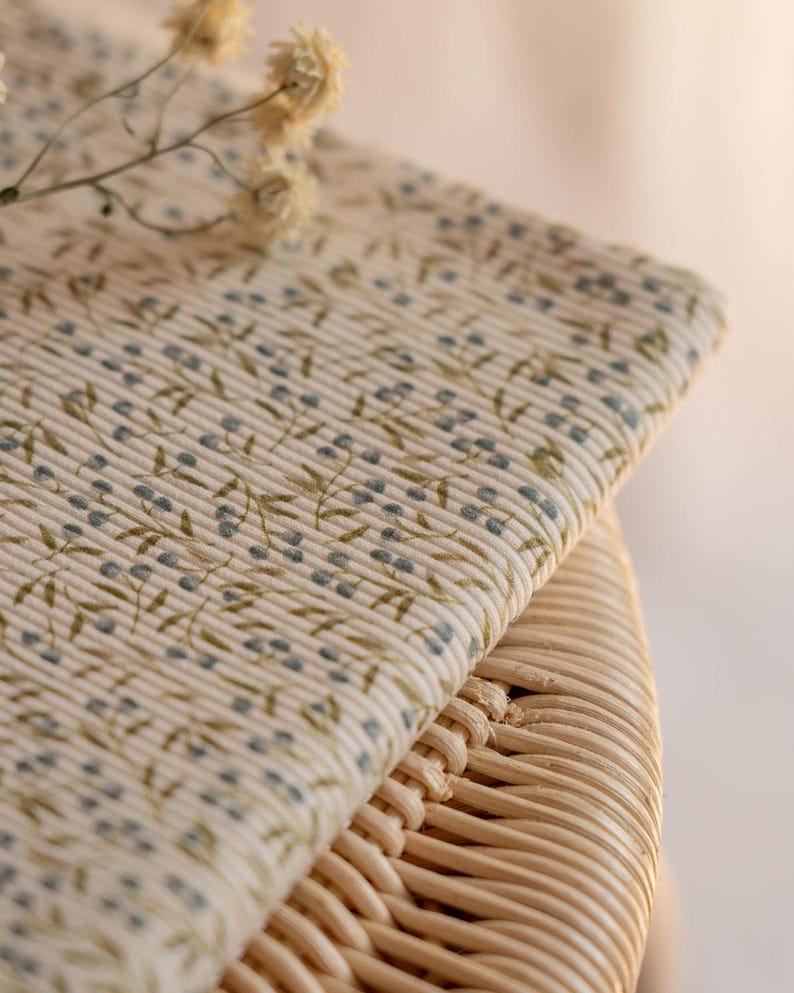 May include: Close-up of folded cream fabric with a floral design. The pattern shows small blue berries and green leaves on stems. The fabric rests on a woven wicker surface. The overall aesthetic is soft and natural.