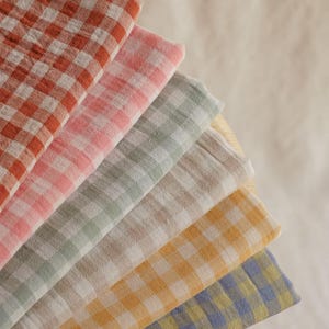 May include: A fan of six folded, soft, checkered fabric cloths in shades of red, pink, green, beige, yellow, and blue. The cloths are arranged in a semi-circle, showcasing the different colors and patterns. The fabric appears to be lightweight.