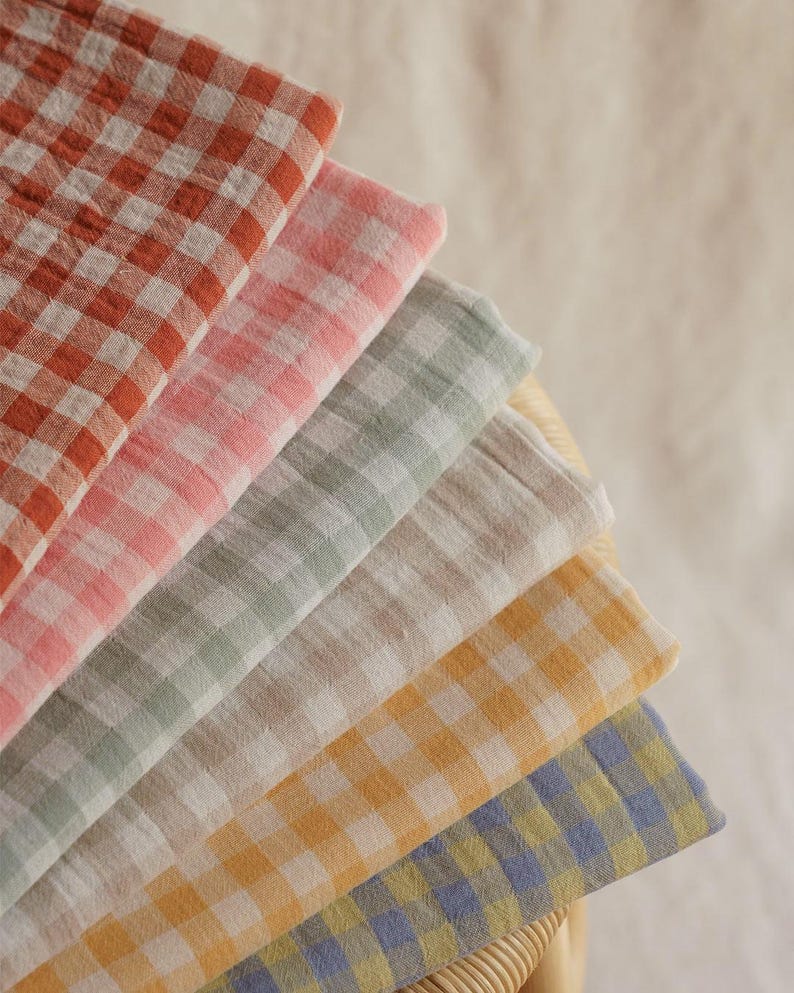 May include: A fan of six folded, soft, checkered fabric cloths in shades of red, pink, green, beige, yellow, and blue. The cloths are arranged in a semi-circle, showcasing the different colors and patterns. The fabric appears to be lightweight.
