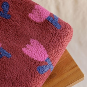 May include: A burgundy fleece blanket featuring a repeating pattern of pink and blue tulip-like flowers. The blanket is folded over a wooden stool, showcasing its soft, textured surface. The floral design adds a touch of whimsy.