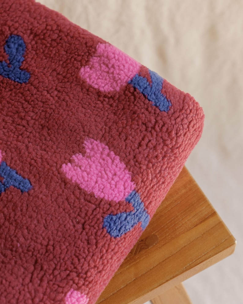 May include: A burgundy fleece blanket featuring a repeating pattern of pink and blue tulip-like flowers. The blanket is folded over a wooden stool, showcasing its soft, textured surface. The floral design adds a touch of whimsy.