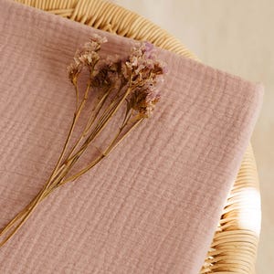 Op de afbeelding: Een gevouwen stuk stof in de kleur dusty rose, mogelijk mousseline of gaas, met een getextureerd oppervlak. Gedroogde, lichtpaarse bloemen liggen bovenop de stof. De stof is geplaatst op een geweven rieten oppervlak.