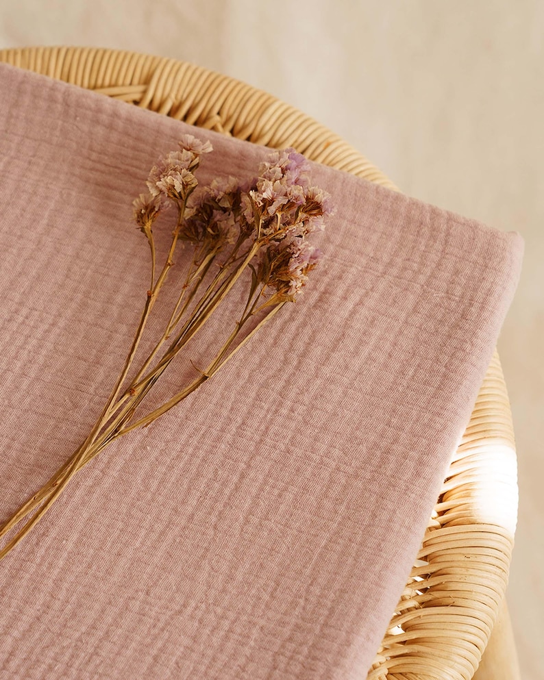 Op de afbeelding: Een gevouwen stuk stof in de kleur dusty rose, mogelijk mousseline of gaas, met een getextureerd oppervlak. Gedroogde, lichtpaarse bloemen liggen bovenop de stof. De stof is geplaatst op een geweven rieten oppervlak.