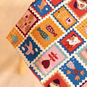 Puede incluir: Una tela con un patrón repetitivo de coloridos diseños de sellos postales. Cada sello cuadrado muestra ilustraciones que incluyen perros, gatos, pájaros, peces, cafeteras, pasteles y corazones en tonos rojos, azules, amarillos y rosas.