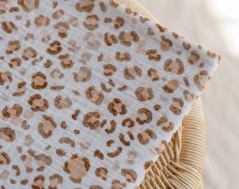 May include: A white fabric with a brown leopard print pattern. The fabric is folded over a wicker chair. The leopard spots are various sizes and shades of brown.