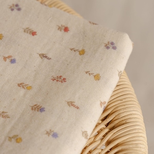 May include: A folded piece of off-white fabric with a delicate floral pattern in shades of red, yellow, and purple. The fabric is resting on a woven wicker surface, creating a contrast in textures. The floral design adds a touch of nature.