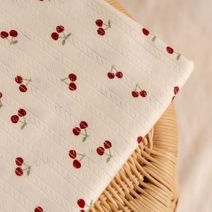 May include: A folded piece of off-white fabric featuring a repeating pattern of small red cherries with green stems. The textile has a subtle vertical ribbed texture with delicate pointelle-style detailing between the rows of cherry motifs.