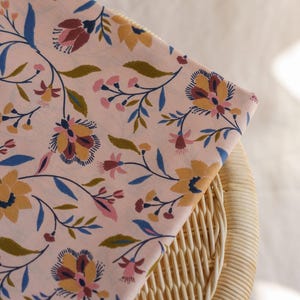 May include: A light pink fabric featuring a repeating floral pattern with stylized flowers and leaves in shades of mustard yellow, deep blue, olive green, and muted rose. The textile displays thin, dark stems connecting the various botanical elements.