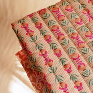 Può includere: Tessuto trapuntato piegato con motivo floreale. Il design presenta file di fiori stilizzati nei toni del pesca e del rosa, con foglie verdi, su uno sfondo crema a sottili strisce verticali. Il tessuto è piegato, rivelando uno scorcio di un motivo complementare.