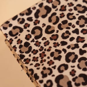 May include: A fabric swatch featuring a repeating leopard print pattern. The design consists of black, dark brown, and tan spots arranged on a cream-coloured background. The material has a visible woven texture and a slightly angled perspective.