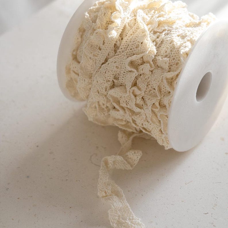 May include: A spool of white crocheted lace trim with a ruffled edge. The lace is wound around a white plastic spool.