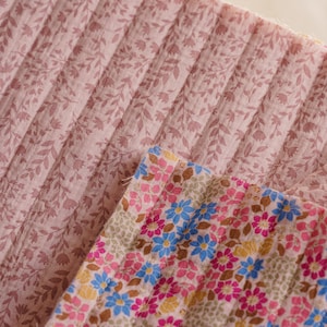 May include: A pleated fabric textile featuring two distinct patterns. The larger section displays a muted mauve background with a delicate, repeating dark pink leaf and vine print. The smaller section shows a vibrant floral pattern with blue, pink, and yellow flowers on a light background.