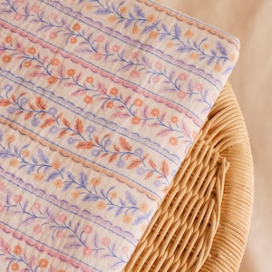 May include: Folded fabric with a floral pattern in shades of peach, lavender, and blue. The design features rows of flowers and leaves, with scalloped edges and blue stripes. The fabric is resting on a woven wicker surface.