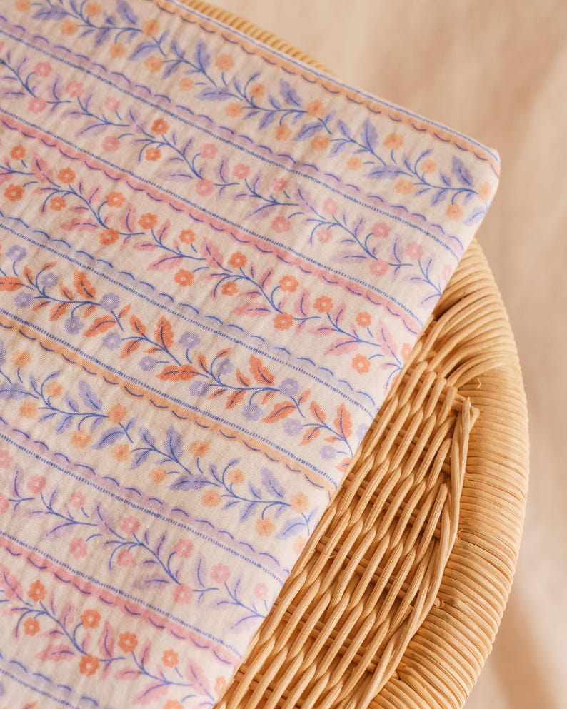 May include: Folded fabric with a floral pattern in shades of peach, lavender, and blue. The design features rows of flowers and leaves, with scalloped edges and blue stripes. The fabric is resting on a woven wicker surface.