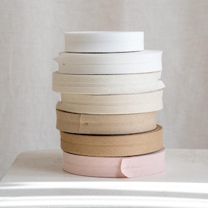 May include: A stack of eight different coloured fabric ribbon trims. The trims are in various shades of white, beige, brown, and pink. The trims are all folded and stacked on top of each other.