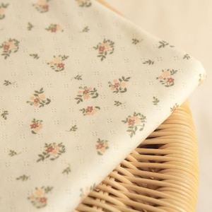 May include: A folded piece of off-white fabric featuring a repeating pattern of small, delicate floral clusters in shades of pink, peach, and green, with a subtle, textured diamond-shaped grid pattern woven into the material surface.