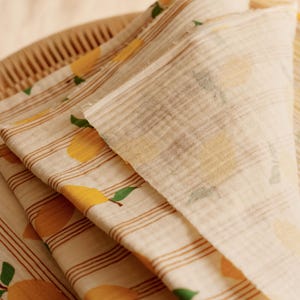 May include: Close-up of folded fabric with a lemon pattern. The fabric is off-white with brown stripes and yellow lemons with green leaves. The fabric is folded over a woven basket.