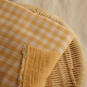 May include: Close-up of a folded fabric with a yellow and white gingham pattern. The fabric is resting on a woven wicker surface. The reverse side of the fabric is a solid yellow color, with a frayed edge.